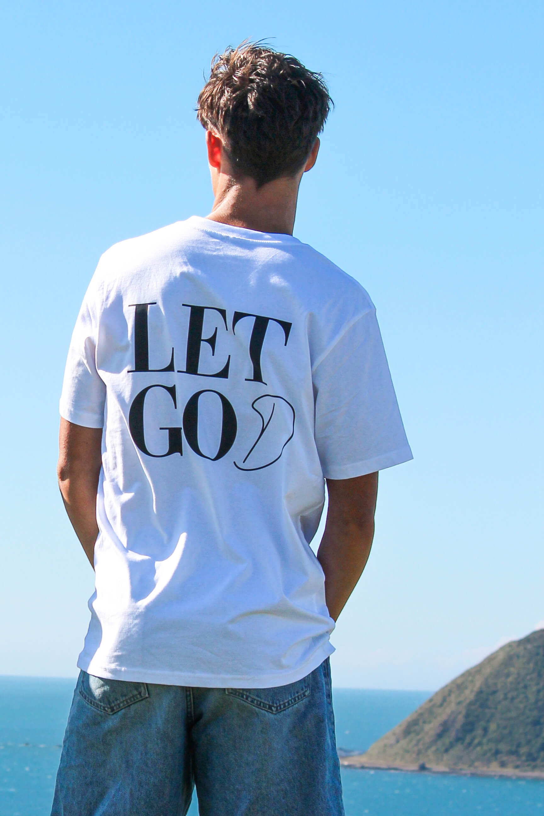 Person wearing a white t-shirt with Sons and Daughters 'LET GO LET GOD' printed on the back, standing against a clear blue sky.